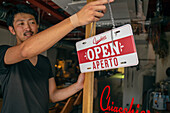 Ein Mann hängt ein Schild "Open" an die Tür eines Restaurants. Zweisprachig, Italienisch und Englisch. 