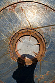 The shadow of a person with arms raised thrown onto the rusted marked surface of a large container with round opening. 