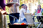Young man preparing food in barbecue grill with his friends sitting at table in background