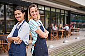 Photo of two happy female restaurant owners standing back to back in front of their restaurant