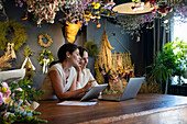 Wide view of two diverse female home decoration shop owners working with laptop computer and digital tablet
