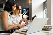 Side view shot of two female interior designers at their desk working with books and a laptop computer