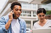 Mid-shot of African-American man speaking on mobile phone and Latin-American woman working on laptop computer outdoors