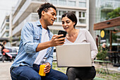 Front view shot of diverse couple hanging out and having fun with their smartphones and computer in an outdoor setting