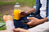 Mid-shot of man's hand holding an orange juice bottle and an out-of-focus hand holding a sandwich