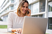 Porträt einer Frau, die im Freien an einem Laptop-Computer arbeitet und dabei mit einem Mobiltelefon telefoniert