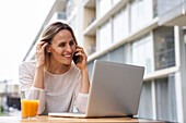 Porträt einer lächelnden, attraktiven Frau, die im Freien sitzt und auf den Bildschirm eines Laptops schaut, während sie mit einem Mobiltelefon spricht und Orangensaft trinkt