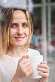 Porträt einer Frau mit Kaffeebecher in der Hand, die durch ein Fenster blickt
