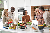 Gruppe von verschiedenen Freunden mittleren Alters, die sich zum Abendessen treffen und in der Küche beim Kochen plaudern