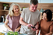 Ein älterer Mann benutzt ein Smartphone, während er sich mit Freunden in der Küche unterhält