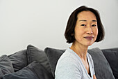 Porträt einer älteren asiatisch-amerikanischen Frau, die in die Kamera schaut und im Wohnzimmer sitzt