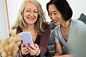 Porträt von zwei Freundinnen mittleren Alters, die im Wohnzimmer auf den Bildschirm eines Mobiltelefons schauen