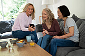 Mittelaufnahme einer Frau mittleren Alters, die zwei Freundinnen Fotos auf ihrem Handy zeigt, während sie zu Hause auf dem Sofa sitzen