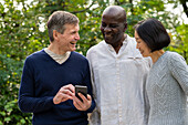 Porträt von drei Freunden, die auf den Bildschirm ihres Mobiltelefons schauen, während sie draußen in einem öffentlichen Park abhängen