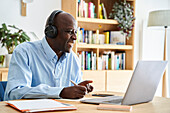 Afroamerikanischer Mann mittleren Alters, der Kopfhörer trägt und sich Notizen macht, während er an einer Fernkonferenz teilnimmt, während er zu Hause mit einem Laptop-Computer arbeitet