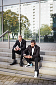 Businessmen sitting on stairs and using phones outside office building