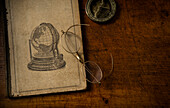 Old book resting on old wooden desk top with old eyeglasses and small sundial