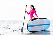 Woman in pink swimsuit standing with paddleboard by lake