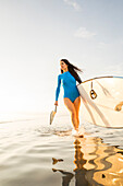 Woman in blue swimsuit carrying paddleboard in lake at sunset