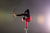 Young acrobat performing with archery bow on gymnastics bar