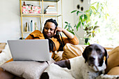 Frau liegt mit Hund auf dem Sofa und arbeitet an einem Laptop