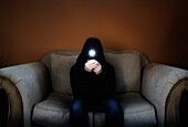 Man in hood sitting on sofa and holding flashlight in front of his face