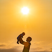 Profile of father holding baby son (6-11 months) aloft against yellow sky at sunset