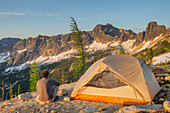Adult man at daybreak sitting next to backpacking tent and gazing at view of Cutthroat Peak on ridge above Cutthroat Pass, near Pacific Crest trail. North Cascades, Washington State