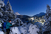 One man photographing the old Chiesa Bianca at night standing in a snowy forest, Maloja, Engadine, Canton of Graubunden, Switzerland, Europe