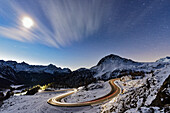 Full moon glowing over a snowy mountain road lit by car trails, Bernina Pass, Val Poschiavo, Graubunden canton, Switzerland, Europe