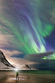 Eine Person genießt die Magie des arktischen Himmels mit den Nordlichtern am Strand von Haukland, Insel Lofoten, Norwegen, Europa