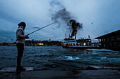 Man fishing in the Golden Horn in Galata neighbourhood while a ferry boat is sailing