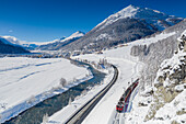 Bernina Express train traveling in the snowy landscape on shore of Inn river, Madulain, Graubunden, Engadin, Switzerland
