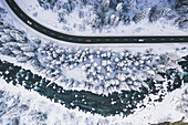 Luftaufnahme von Autos, die im verschneiten Wald am Rande eines zugefrorenen Flusses im Winter unterwegs sind, Schweiz