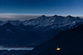 Sternennacht über beleuchteter Kapelle und schneebedeckten Bergen des Alto Lario, Costiera dei Cech, Valtellina, Lombardei, Italien