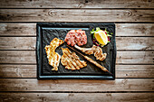 Mixed grilled meat in black tray on rustic wood table background from above, Italy