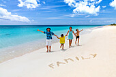 Glückliche Familie mit zwei kleinen Jungen, die in die Kamera lächeln, neben dem auf Sand geschriebenen Wort Familie, Antigua und Barbuda, Karibik