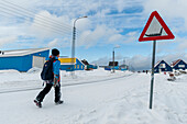 A kid walking home on a snow-covered street passing a dog sled crossing warning sign. Ilulissat, Greenland.