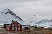 A plane flies above multicolored buildings and mountains at Ny-Alesund. Ny-Alesund, Kongsfjorden, Spitsbergen Island, Svalbard, Norway.