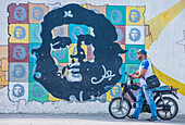 A man with a motorcycle walking past a mural of Che Guevara in old Havana street. The historic center of Havana is UNESCO World Heritage Site since 1982.