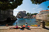 Menschen entspannen sich am Strand von Dubrovnik, gegenüber dem Fort Lovrijenac, Dubrovnic, Kroatien. Dies ist ein Foto von Menschen, die sich an einem kleinen Strand in Dubrovnik, gegenüber dem Fort Lovrijenac, Kroatien, entspannen. Das Fort Lovrijenac ist eine große Festung, die auf einem Felsen liegt, gleich außerhalb der Altstadt von Dubrovnik.