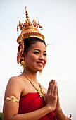 Young Thai woman in traditional costume with golden crown and jewelry and hands together in traditional "wai" greeting; at Siam Niramit, Bangkok, Thailand.