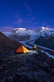 Wanderer mit Stirnlampe neben dem Zelt sitzend vor Monte Rosa Massiv, Gornergletscher und Lyskammspitzen in der Abenddämmerung, Riffelsee, Zermatt, Kanton Wallis, Schweizer Alpen, Schweiz, Europa
