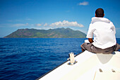 Man Sitting On Bow Of Boat Traveling Towards Island