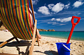 Frau auf Liegestuhl neben Plastikeimer und Spaten am Strand von Porthcurno