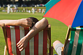 Rückansicht von Zuschauern in Liegestühlen, die ein Kricketspiel an einem heißen Sommertag in Hastings verfolgen.
