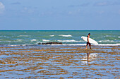 Mann trägt Surfbrett am Praia Do Forte, Bahia, Brasilien