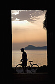 Silhouette eines Mannes, der in der Abenddämmerung ein Fahrrad entlang der Strandpromenade schiebt, Seitenansicht, Lopud, Kroatien