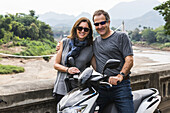Ein Paar posiert neben einem Motorrad mit einer bergigen Landschaft in der Ferne; Luang Prabang, Provinz Luang Prabang, Laos.