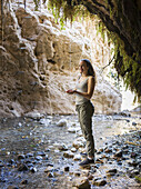 Eine junge Frau steht auf einem Felsen entlang eines Baches in der Dodim-Höhle, Naturreservat Ein Gedi; Südregion, Israel.
